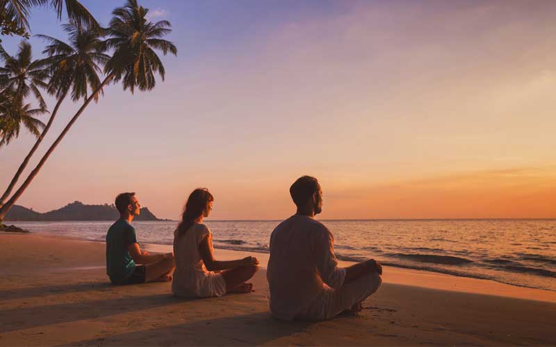 3 people meditating on a beach looking out to the water